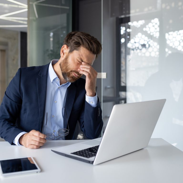 Stressed businessman with hand on forehead sitting at desk with laptop, tablet, and paperwork in modern office.